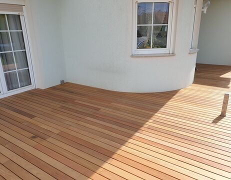 Neu installierte Holzterrasse neben einem Haus mit weißen Wänden, zwei Fenstern und einer Glastür.