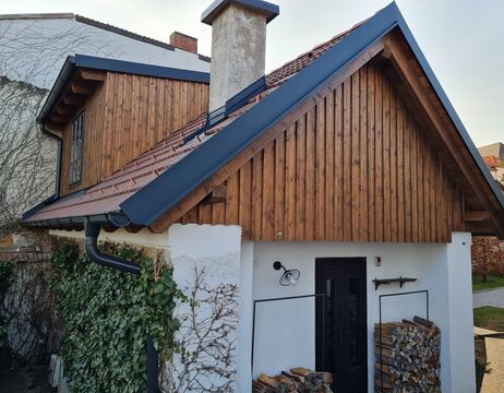 Kleines Haus mit holzgetäfelten oberen Wänden, gestapeltem Brennholz und Efeu, der an den weißen unteren Wänden wächst.