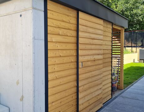 Moderner Schuppen mit heller Holzverkleidung, Betonwand und Schiebetüren neben einem grasbewachsenen Hof.