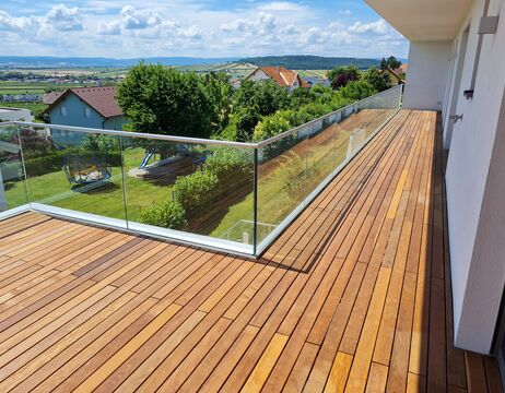 Moderner Holzbalkon mit Glasgeländer mit Blick auf einen grünen Hof und ferne Hügel unter blauem Himmel.
