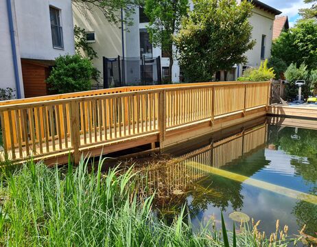 Eine Holzbrücke mit Geländer führt über einen kleinen Teich, der von modernen Häusern und Grünanlagen umgeben ist.