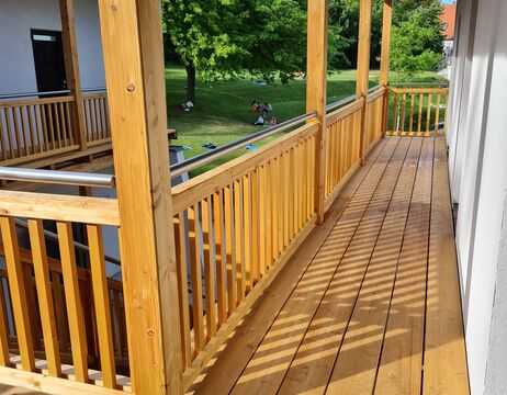 Ein hölzerner Balkongang mit Geländer blickt auf eine Rasenfläche, auf der Menschen unter einem Baum sitzen.