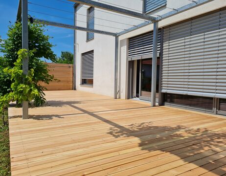 Modernes Haus mit großen Fenstern und Metallpergola auf einer großen Holzterrasse, sonniger Tag mit Baumschatten.