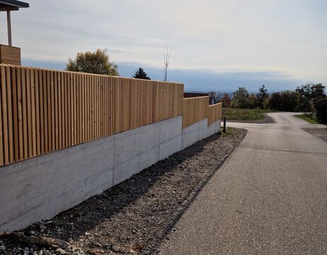 Ein moderner Zaun aus Holz und Beton säumt eine ruhige, kurvenreiche Straße unter einem teilweise bewölkten Himmel.