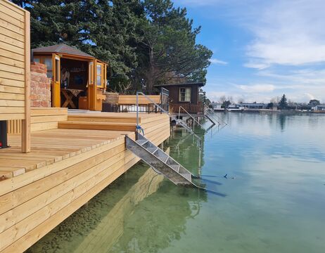 Holzdeck mit Stufen, die ins klare Wasser des Sees führen, kleine Hütte und Bäume im Hintergrund unter blauem Himmel.