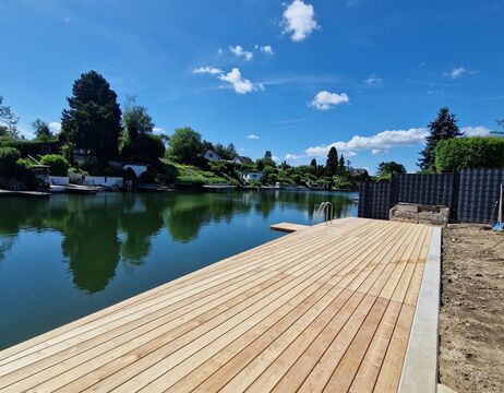 Hölzerner Steg an einem ruhigen Fluss mit Bäumen, Häusern und blauem Himmel mit Wolken im Hintergrund.