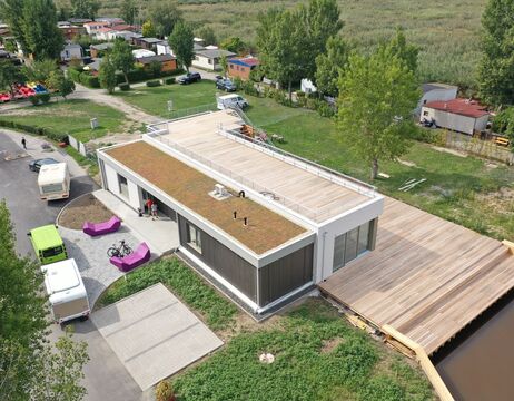 Luftaufnahme eines modernen Gebäudes mit begrüntem Dach, Holzterrasse und Wohnwagen in der Nähe in einer ländlichen Gegend.