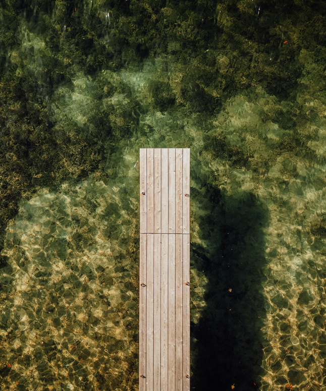 Luftaufnahme eines Holzstegs, der sich über klares, grünliches Wasser mit sichtbaren Mustern auf dem Seegrund erstreckt.