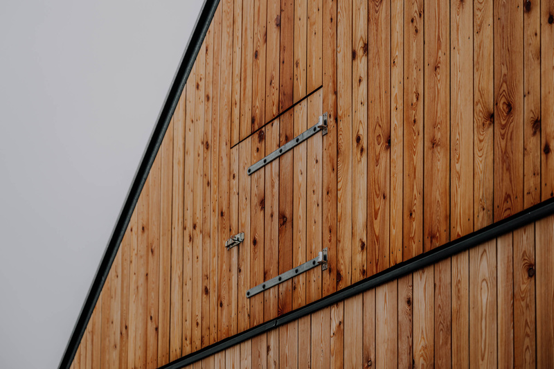 Nahaufnahme einer hölzernen Scheunenwand mit einer kleinen rechteckigen Tür und Metallscharnieren, schräg aufgenommen.