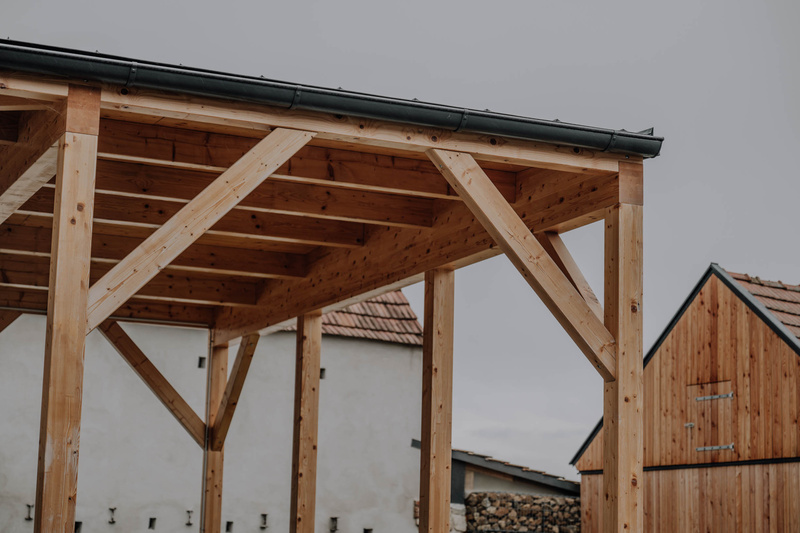 Hölzernes Pergola-Gestell unter bewölktem Himmel, mit Häusern und Holzstapeln im Hintergrund.