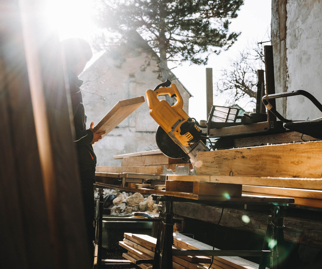 Person hält Holz in der Nähe einer Kreissäge in einer sonnenbeschienenen Werkstatt im Freien mit Bäumen und Gebäuden im Hintergrund.