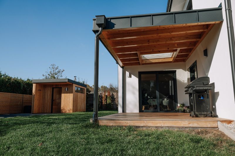 Modernes Haus mit Holzterrasse, Grill und einem kleinen Holzschuppen in einem grasbewachsenen Hinterhof.
