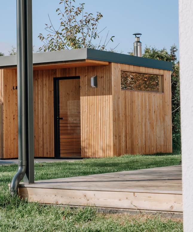 Kleiner moderner Holzschuppen mit Flachdach in einem Hinterhof, umgeben von Gras und einem Holzzaun.