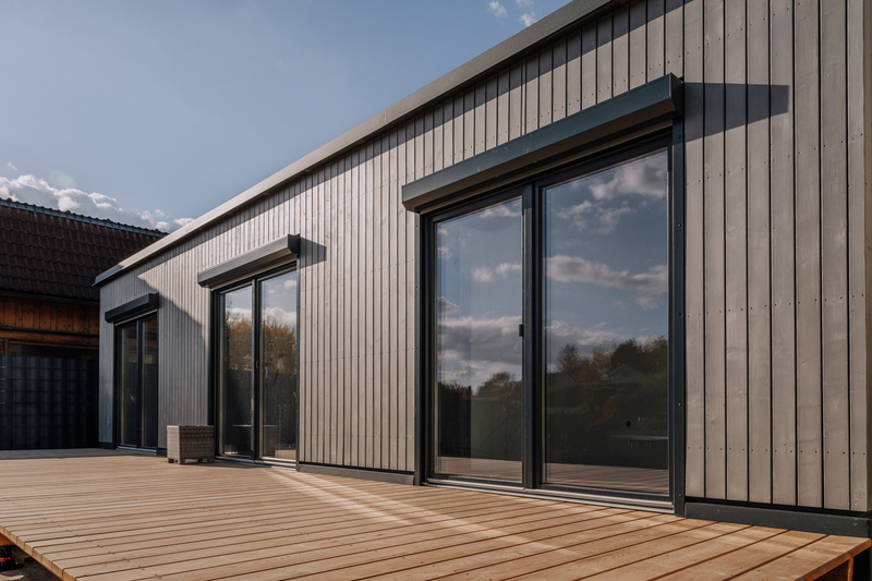 Modernes Holzhaus mit großen Glasschiebetüren und einer großen Holzterrasse unter blauem Himmel.