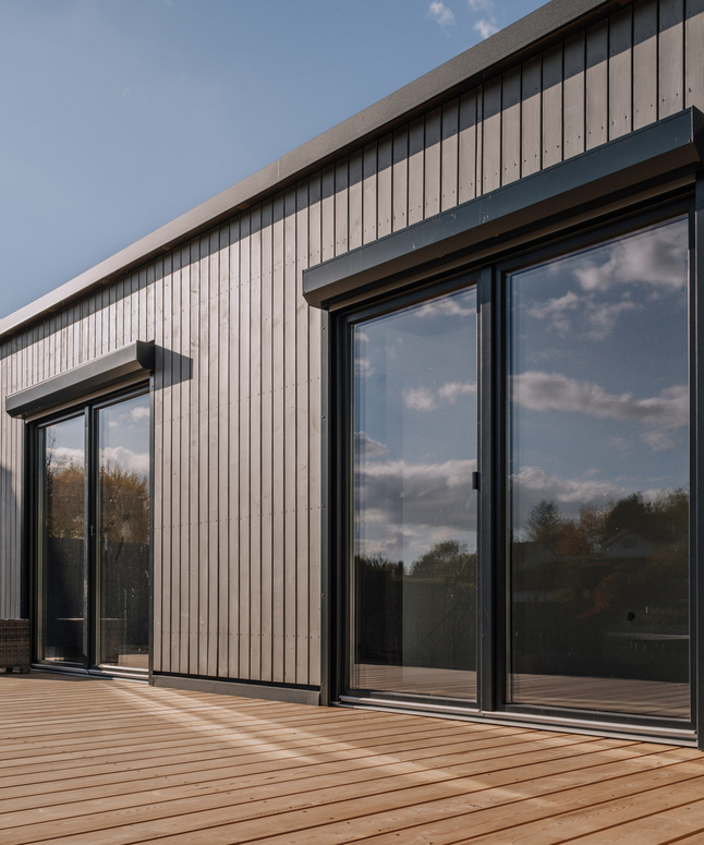 Modernes Holzhaus mit großen Glasschiebetüren und einer großen Holzterrasse unter blauem Himmel.
