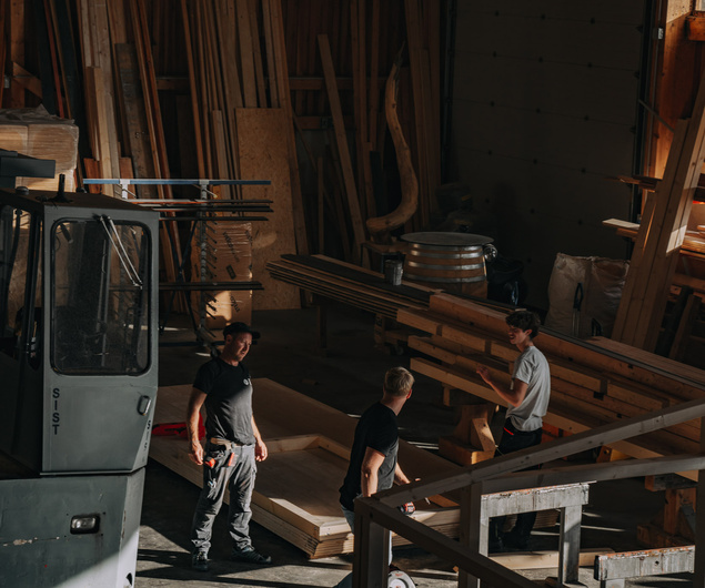 Drei Männer unterhalten sich und arbeiten mit Holzbrettern in einer Werkstatt, die mit Holz und Werkzeugen gefüllt ist.