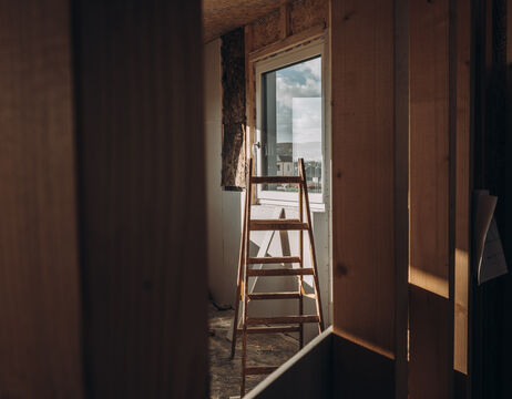 Eine hölzerne Leiter steht in einem teilweise ausgebauten Raum in der Nähe einer offenen Tür, durch die Sonnenlicht einfällt.
