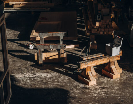 Holzbearbeitungswerkzeuge und Bänke in einer sonnendurchfluteten Werkstatt mit verstreutem Holz und Geräten.