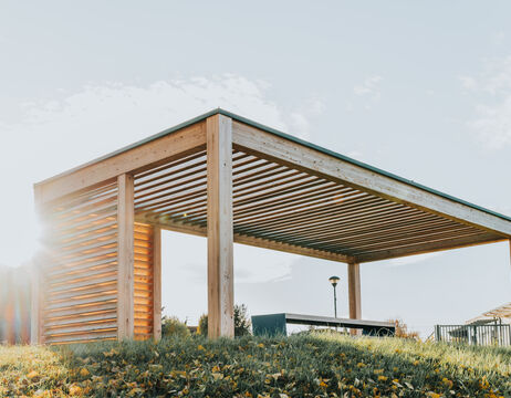 Moderne Holzpergola auf einem Grashügel, bei der das Sonnenlicht durch das Lamellendach und die Seitenwände scheint.