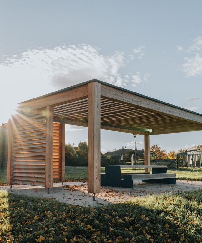 Eine moderne hölzerne Pergola mit Bänken steht auf einer Wiese, das Sonnenlicht scheint durch die Lamellen.