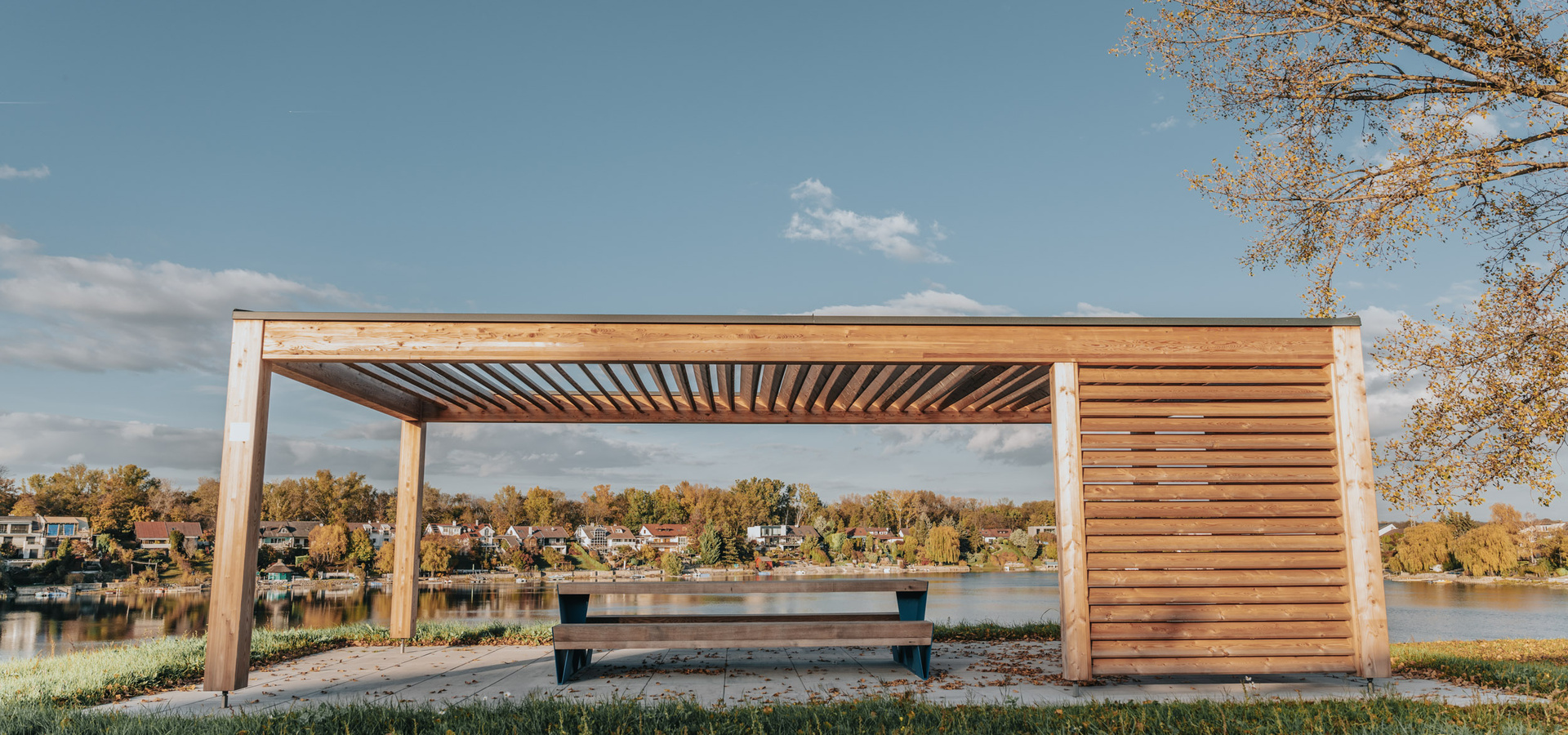 Hölzerne Pergola mit einer blauen Bank darunter, mit Blick auf einen ruhigen See und Häuser im Hintergrund.