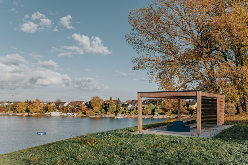 Ein hölzerner Unterstand mit Bänken überblickt einen ruhigen See, mit Bäumen und Häusern im Hintergrund an einem sonnigen Tag.