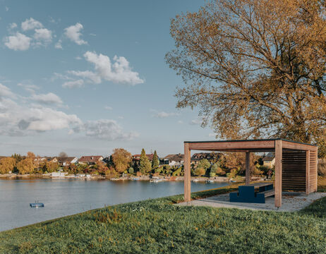 Ein hölzerner Unterstand mit Bänken überblickt einen ruhigen See, mit Bäumen und Häusern im Hintergrund an einem sonnigen Tag.