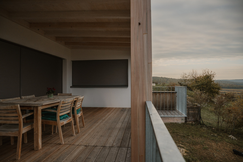 Überdachte Holzveranda mit Esstisch und Stühlen, mit Blick auf eine ländliche Landschaft unter einem bewölkten Himmel.