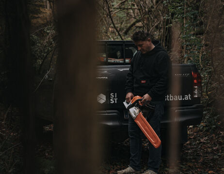 Ein Mann mit einer Kettensäge steht neben einem schwarzen Pickup in einem Waldgebiet, das teilweise von Bäumen verdeckt wird.