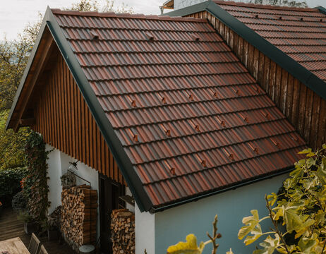 Steiles rotes Ziegeldach auf einem Holzhaus mit gestapeltem Brennholz an der Wand und Grünzeug in der Nähe.