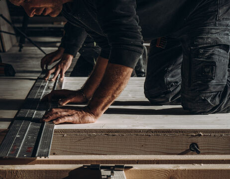 Eine Person, die auf dem Boden kniet, misst mit einem Lineal Holz aus und arbeitet an einem Tischlerprojekt.