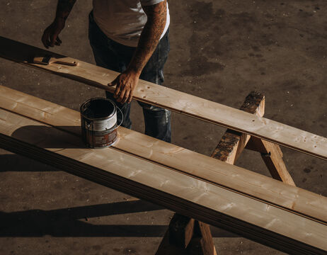 Person beim Beizen von Holzdielen mit einem Pinsel, Dose mit Holzbeize auf der Diele, in einer Werkstattumgebung.
