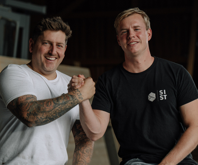 Zwei lächelnde Männer schütteln sich die Hände, der eine in einem weißen Hemd mit Tätowierungen, der andere in einem schwarzen T-Shirt mit einem Logo.