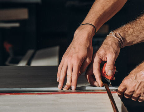 Nahaufnahme von Händen, die ein Holzbrett mit einem Maßband auf einer Werkbank messen.