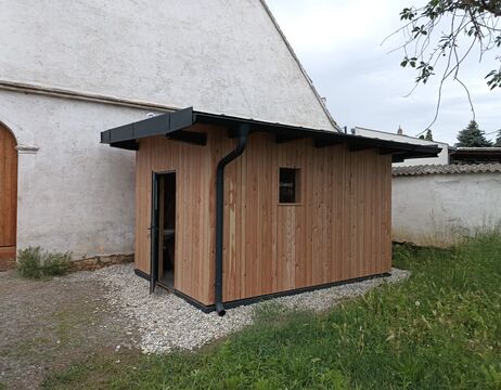 Ein kleiner Holzschuppen mit einem schrägen Dach und einer Tür, neben einem weißen Gebäude, auf Kies und Gras.