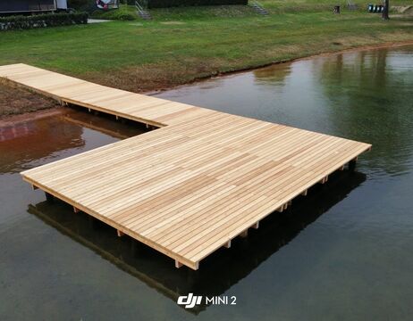 Ein Holzsteg erstreckt sich über ruhiges Wasser in der Nähe eines grasbewachsenen Ufers. Das DJI Mini 2-Logo erscheint am unteren Rand.