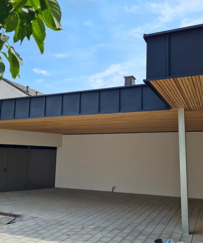 Modernes Carport mit Flachdach, Holzdecke und gefliestem Boden unter blauem Himmel, teilweise Baum sichtbar.