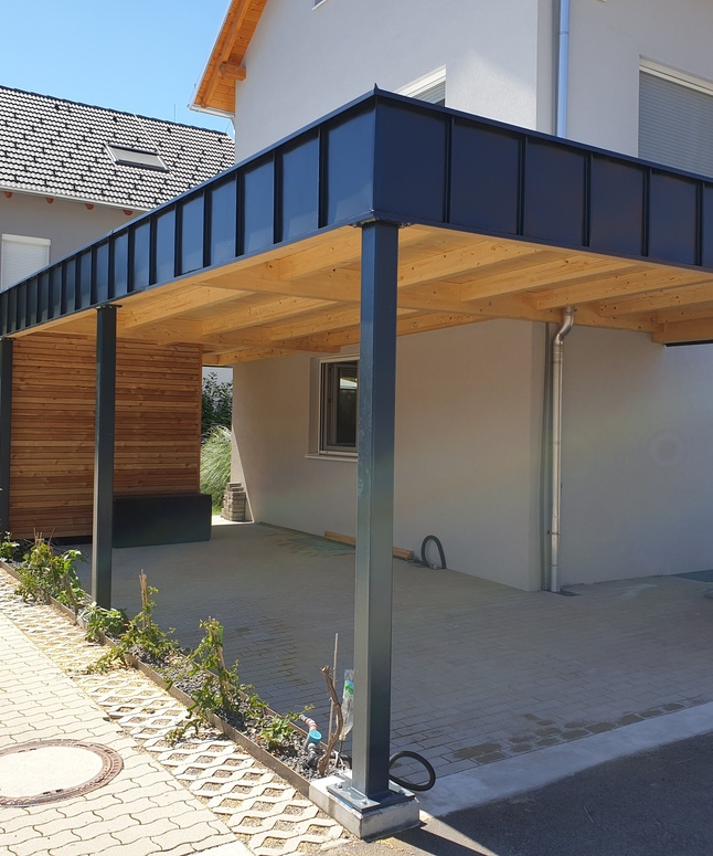 Modernes Carport mit Metalldach und Holzwänden an einem hellgrauen Haus an einem sonnigen Tag.
