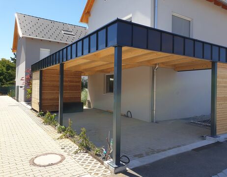 Modernes Carport mit Metalldach und Holzwänden an einem hellgrauen Haus an einem sonnigen Tag.