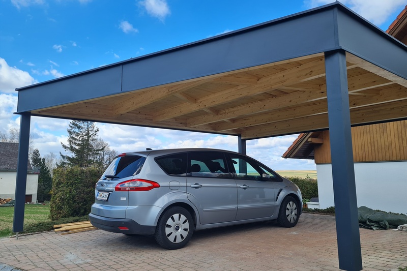 Ein silbernes Auto, das an einem bewölkten Tag unter einem modernen Holzcarport neben einem Haus geparkt ist.