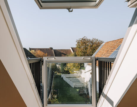 Blick durch ein offenes Dachfenster auf Dächer, einen Garten und einen weißen Lattenzaun an einem klaren Tag.
