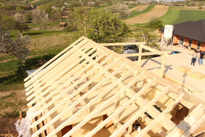 Im Bau befindlicher Dachstuhl aus Holz in einer ländlichen Gegend mit Feldern, Bäumen und einem Haus im Hintergrund.