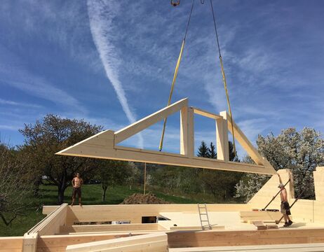 Ein Kran hebt einen großen hölzernen Dachstuhl über ein im Bau befindliches Haus, während zwei Arbeiter ihn an seinen Platz führen.