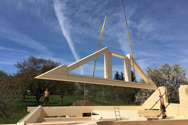 Ein Kran hebt einen großen hölzernen Dachstuhl über ein im Bau befindliches Haus, während zwei Arbeiter ihn an seinen Platz führen.