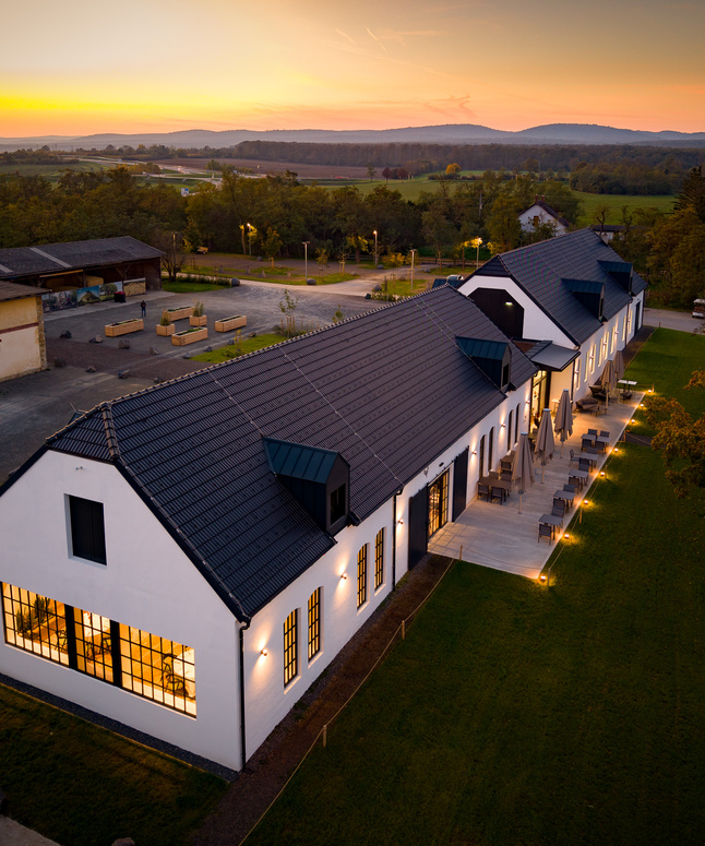 Langes weißes Gebäude mit schwarzem Dach, Außenbeleuchtung und einem Sonnenuntergang im Hintergrund, von oben gesehen.