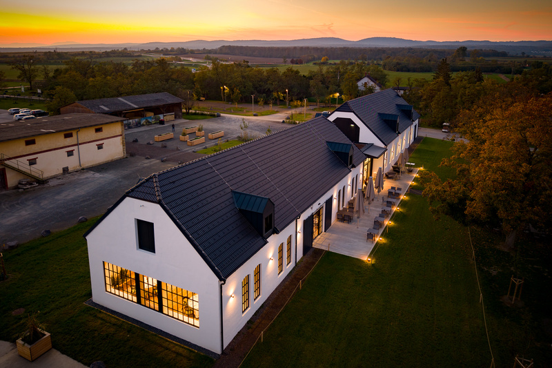 Langes weißes Gebäude mit schwarzem Dach, Außenbeleuchtung und einem Sonnenuntergang im Hintergrund, von oben gesehen.