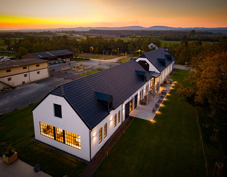 Langes weißes Gebäude mit schwarzem Dach, Außenbeleuchtung und einem Sonnenuntergang im Hintergrund, von oben gesehen.