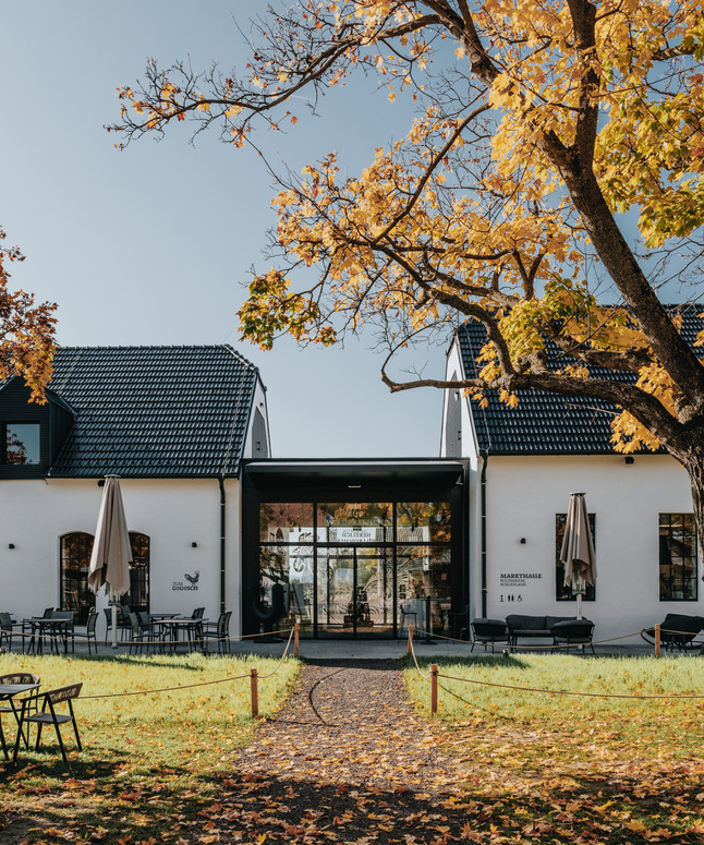 Weißes Gebäude mit schwarzem Dach, Sitzgelegenheiten im Freien und Herbstbäumen mit gelben Blättern davor.