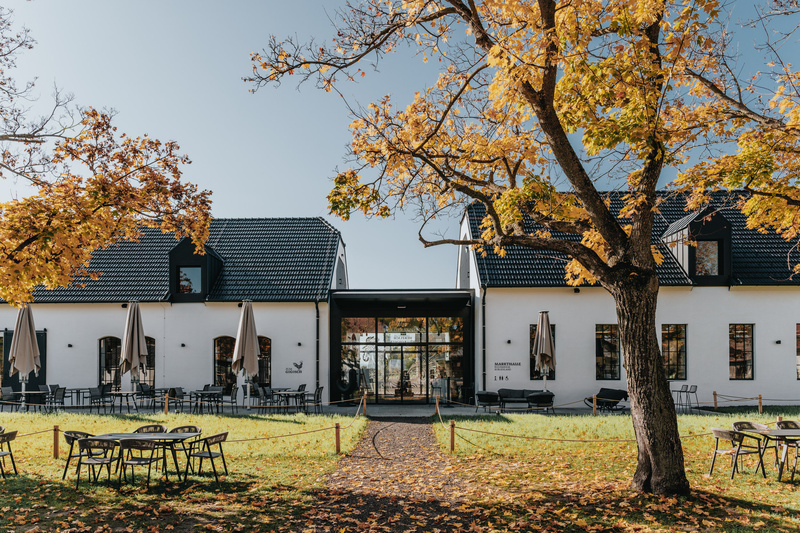 Weißes Gebäude mit schwarzem Dach, Sitzgelegenheiten im Freien und Herbstbäumen mit gelben Blättern davor.