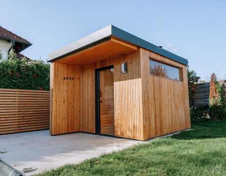 Modernes Gartenhaus aus Holz mit vertikalen Latten, großem Fenster und Flachdach, neben einer Rasenfläche.